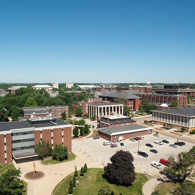 Aerial view of a university campus with brick buildings, pathways, and parking lot under a clear blue sky.