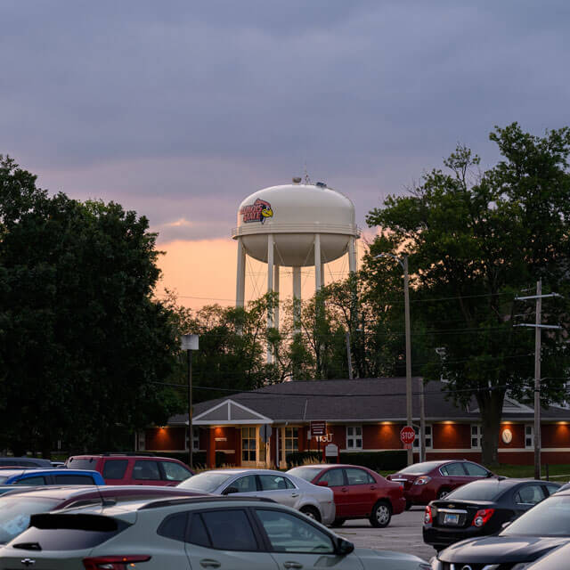 Parking lot with cars, a water tower with 'Illinois State' logo, and trees against a sunset sky.